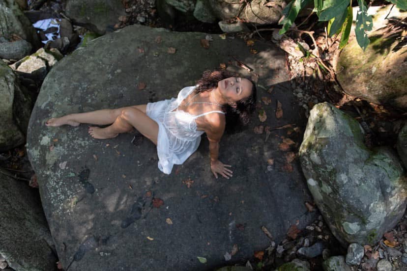 Shakti Elaine in a white dress, reclining on a large stone in the forest with her face turned to the light