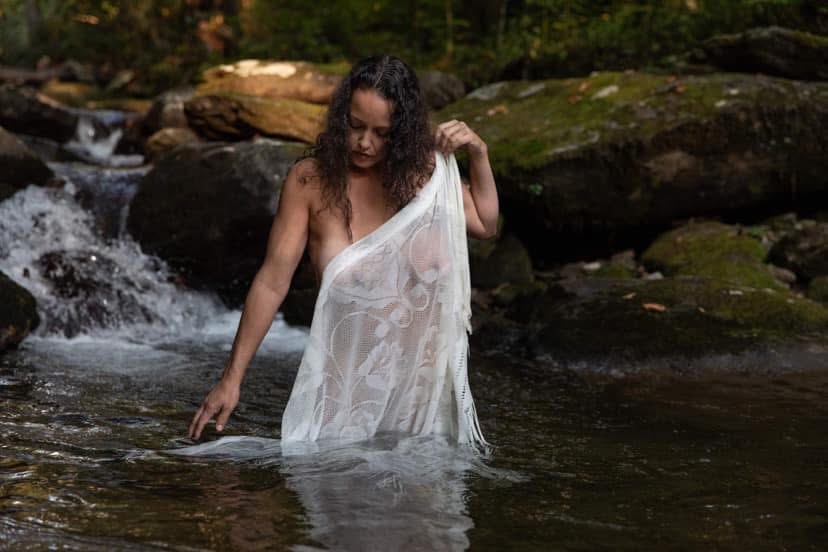 Shakti Elaine standing in a forest stream, draped in white lace